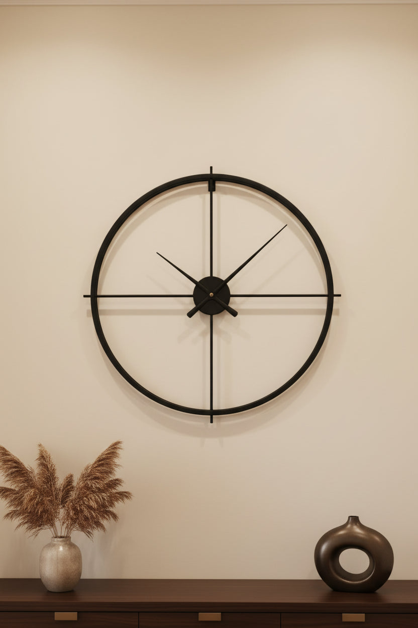 A minimalist black metal wall clock with a large circular frame and cross-shaped hands, mounted on a wall.