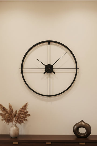 A minimalist black metal wall clock with a large circular frame and cross-shaped hands, mounted on a wall.