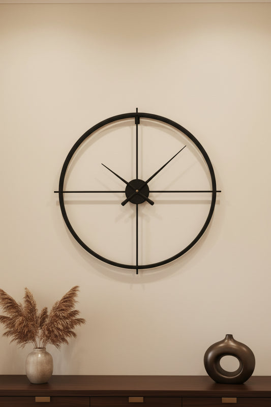 A minimalist black metal wall clock with a large circular frame and cross-shaped hands, mounted on a wall.