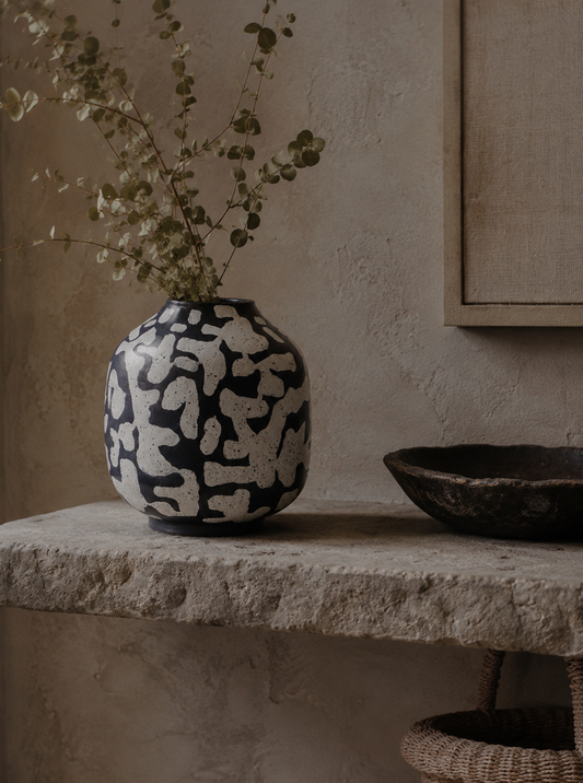 Black and white patterned vase with dried plants on a stone surface against a beige wall.